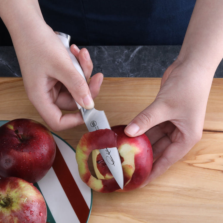 Hands peeling a red apple with a small stainless steel paring knife over a wooden cutting board.