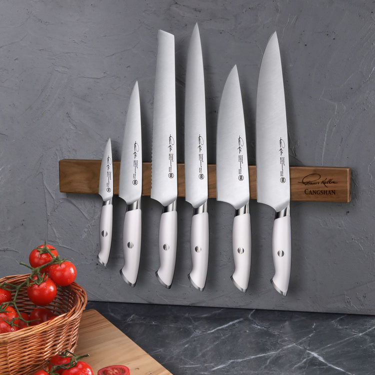 Seven stainless steel knives with white handles mounted on a walnut magnetic knife bar against a gray wall.