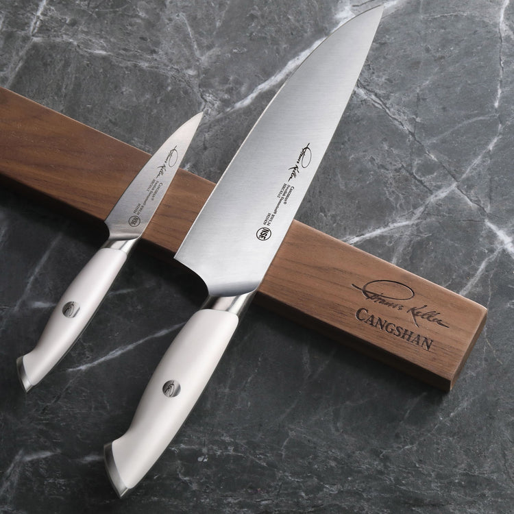 Two stainless steel knives with white handles resting on a walnut magnetic knife bar on a gray marble surface.
